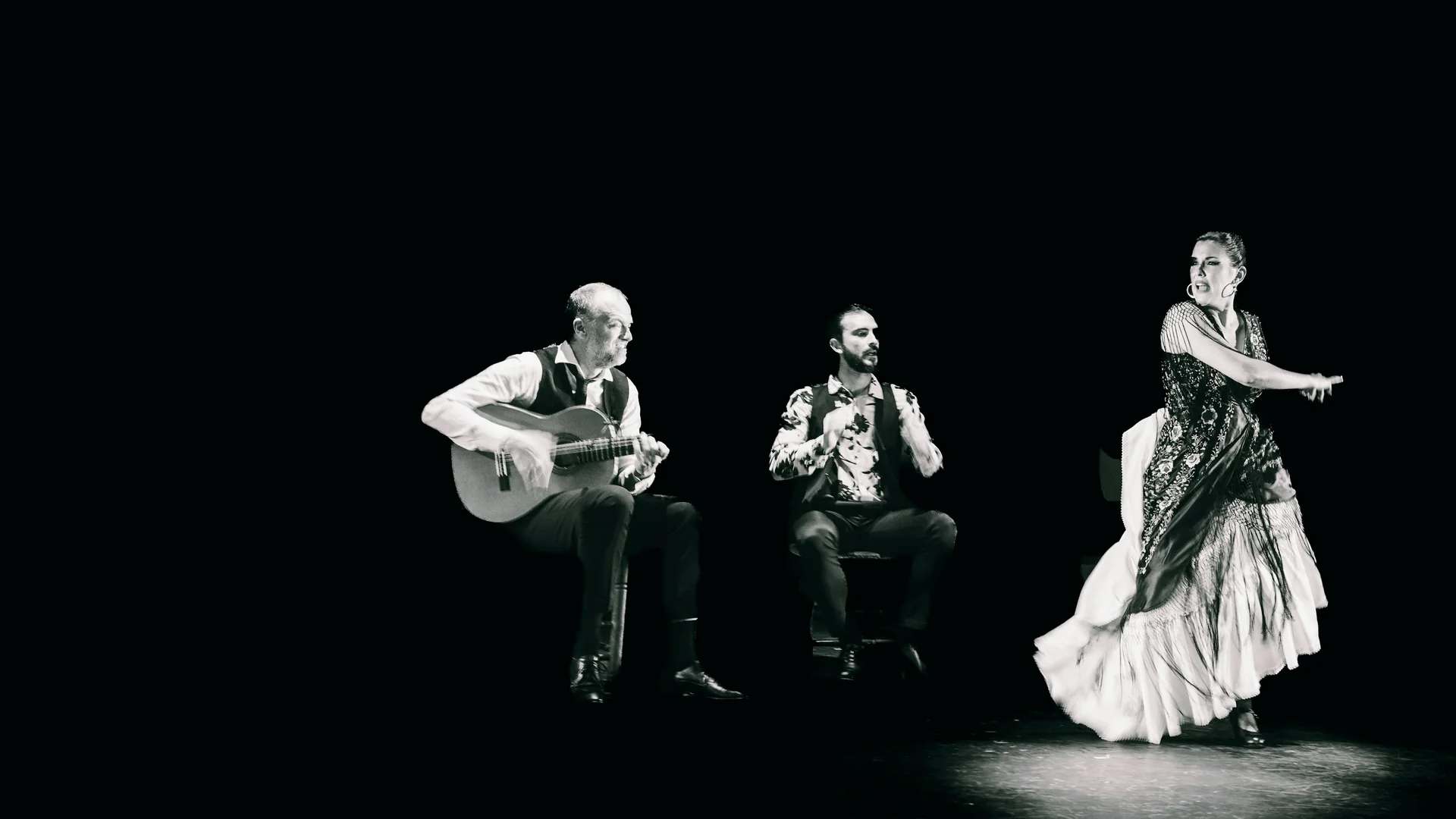 Flamenco tanečnice v bílé sukni s volány a třásnitém šálu v půlobratu vedle sedícího kytaristy a druhého hudebníka, na tmavém jevišti, černobíle